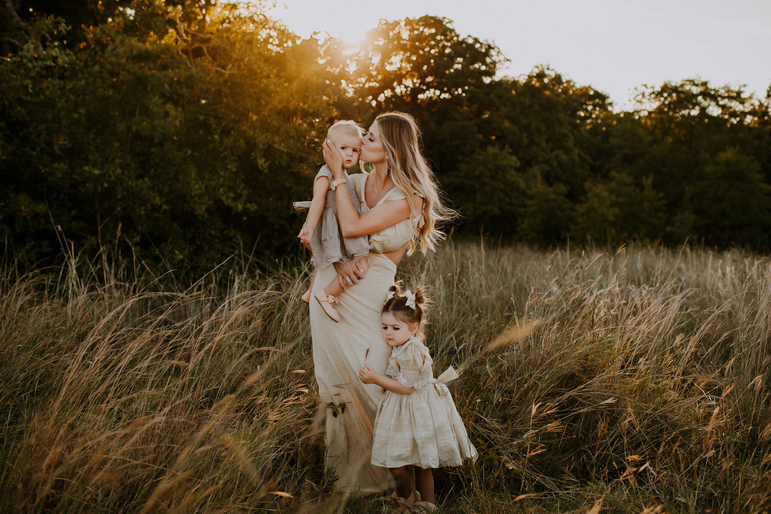 A mom in a cream dress from Revolve standing in a tall grassy field holding one toddler girl and kissing her cheek while her other daughter holds some grass and hugs her leg in fort worth, texas