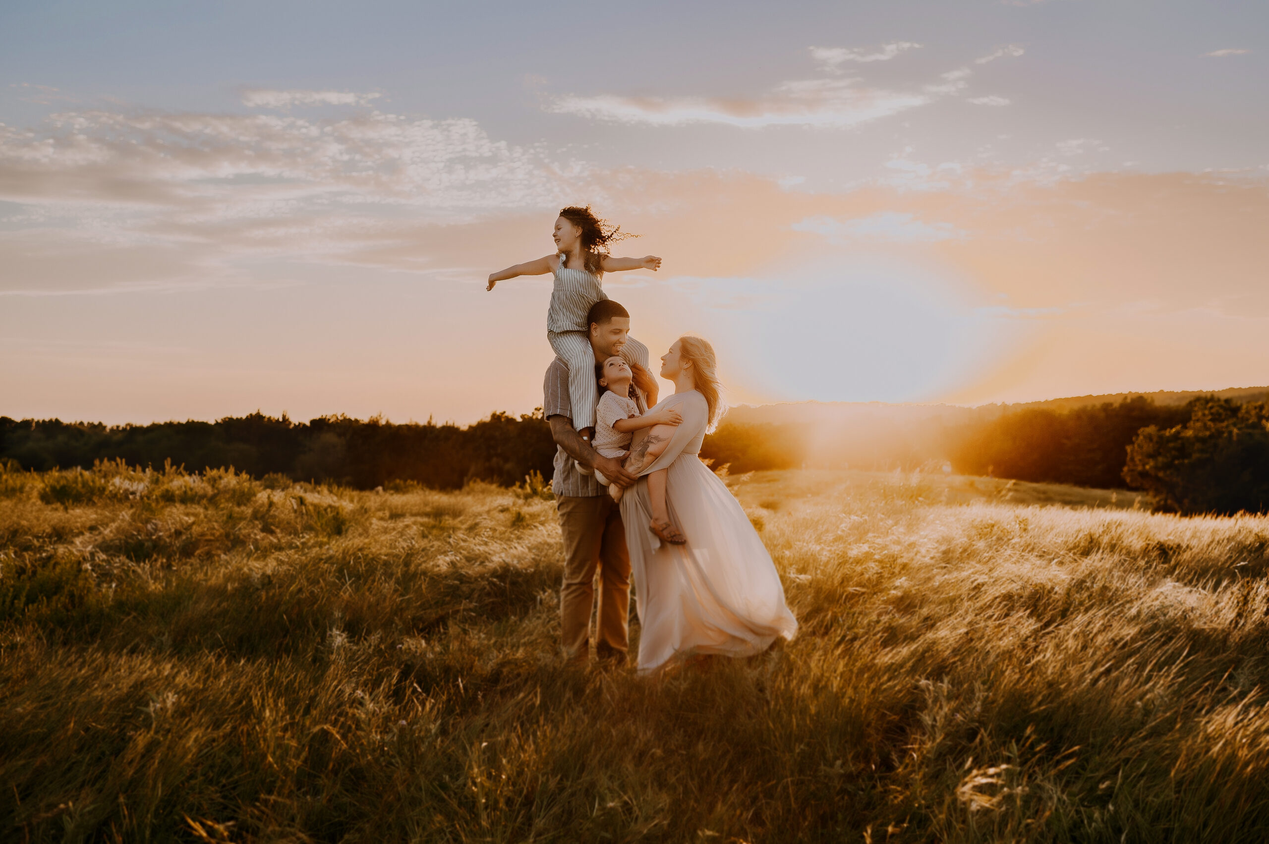 A family of four are standing in the middle of a field at Arbor Hills Nature Preserve in Plano, Texas at sunset in boho fashin cuddling and smiling at one another