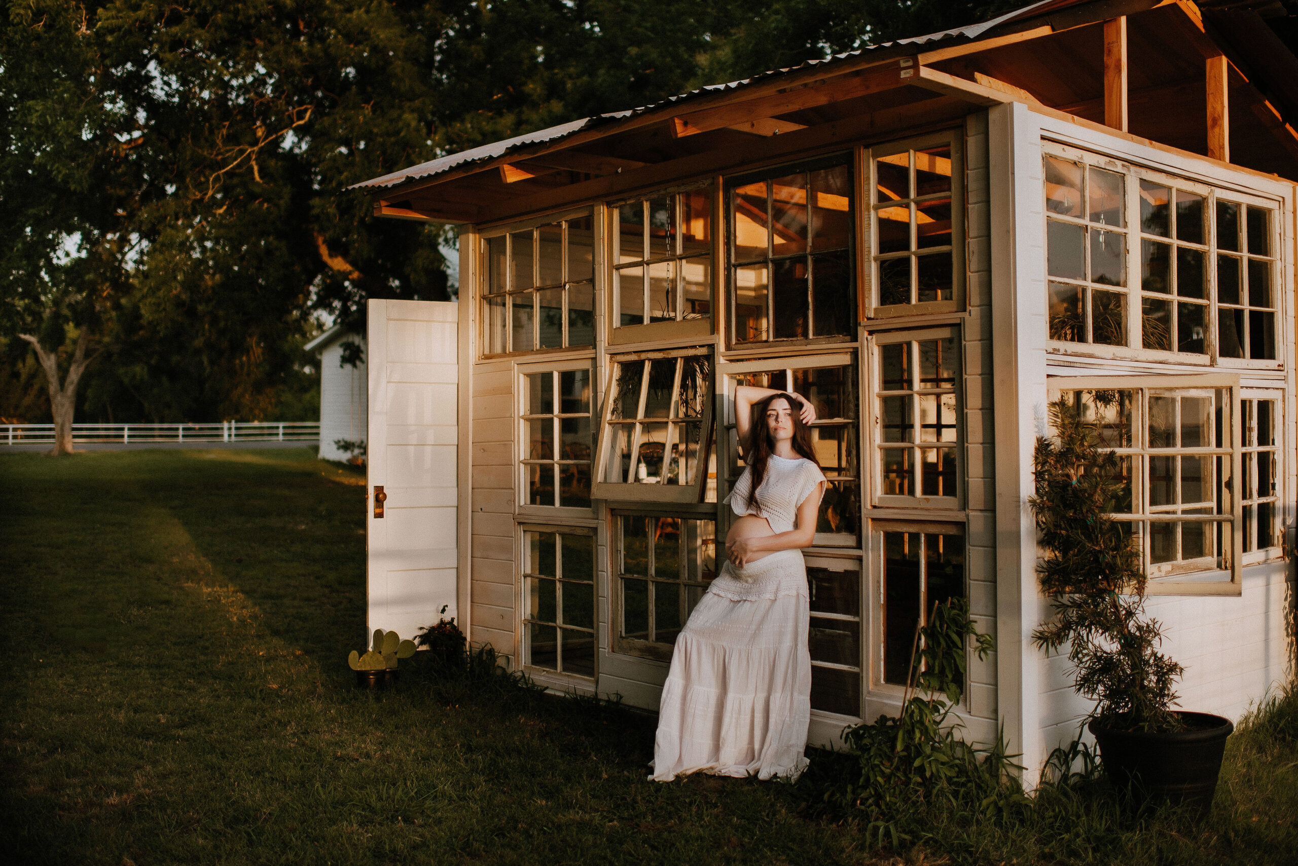 a pregnant woman holding her bare belly with her other arm over her head standing against a glass window plant greenhouse in dallas, texas at the little glass shack