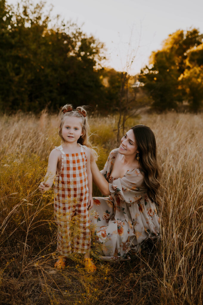 A mom sitting in a tall, grassy field at Northwest Community Park in Fort Worth, Texas holding her daughters shoulder and smiling at her in boho fashion.