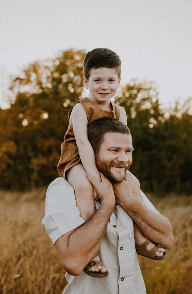 A dad holding his toddler boy on his shoulders while the boy wears overalls from zara and smiles at the camera in fort worth, texas