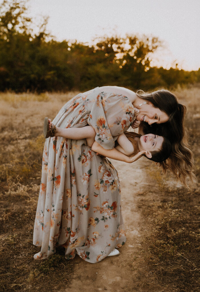 A mom is dipping her laughing son in her arms while wearing a floral dress at Northwest Community Park in Fort Worth, Texas
