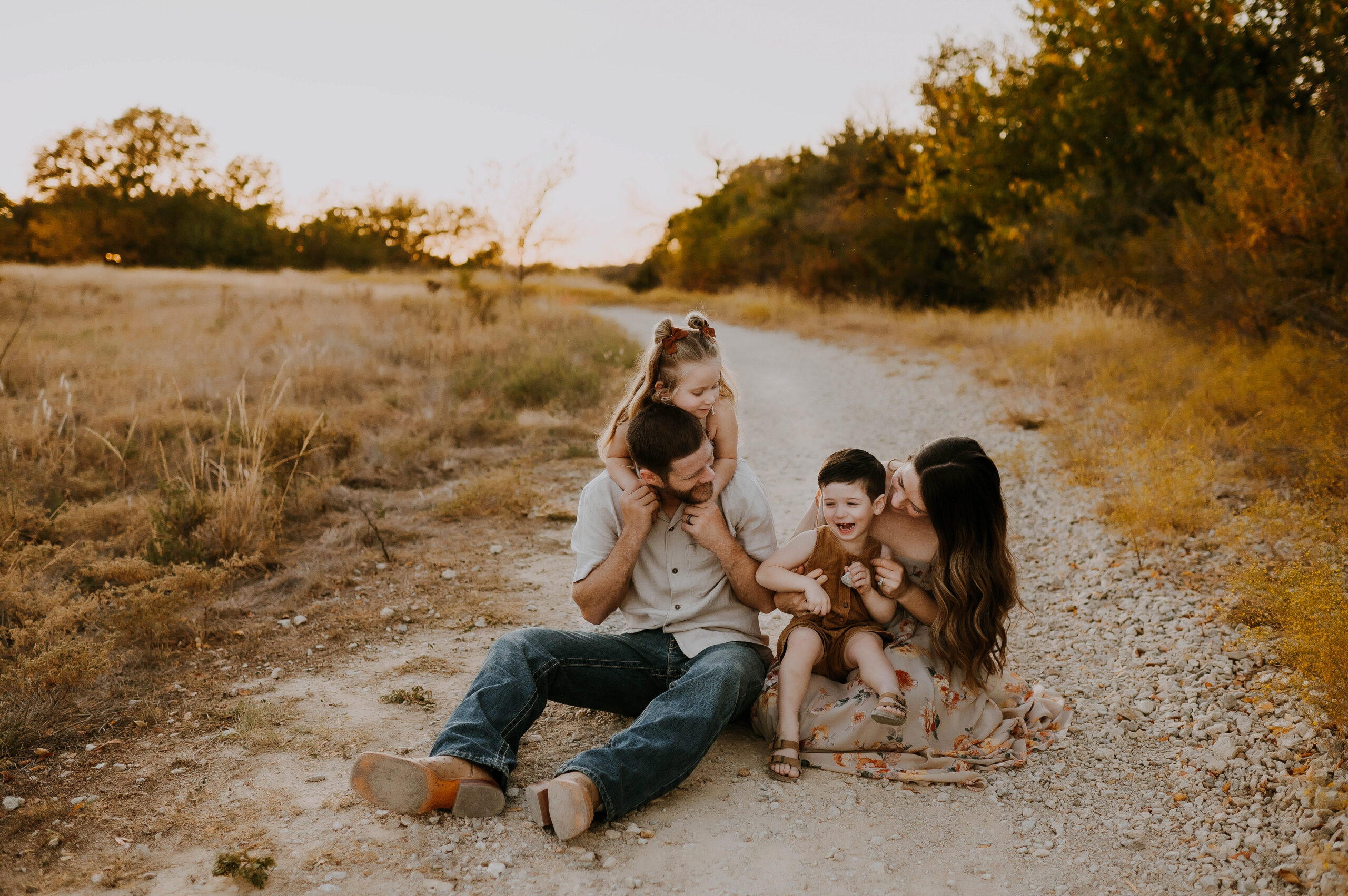 A mom and dad sit on a rocky path in fort worth, texas while their 2 kids laugh and play with them.