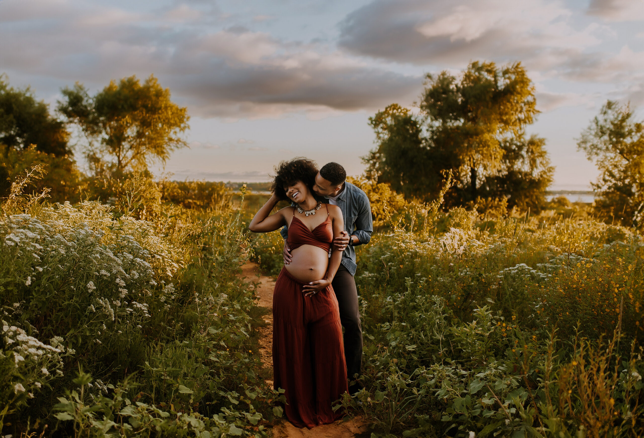 A bare belly pregnant woman in a two-piece set is standing on a trail at Murrell Park in Flower Mound, Texas with her husband as he leans over her shoulder and kisses her neck and she laughs