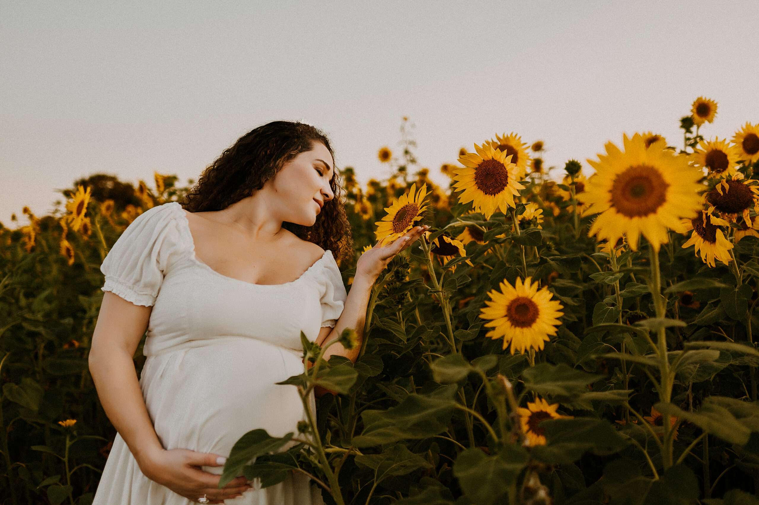 a pregnant woman standing next to a giant sunflower and holding it with her eyes closed in a field in fort worth, texas