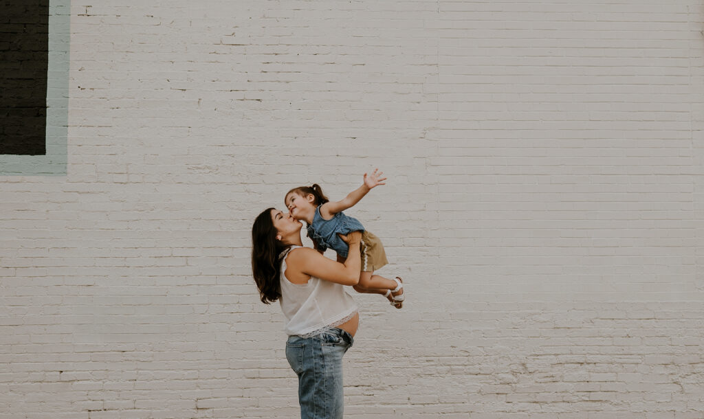 A mom holds her little girl up in the air and kisses her cheek next to an antique mall in downtown mckinney, texas