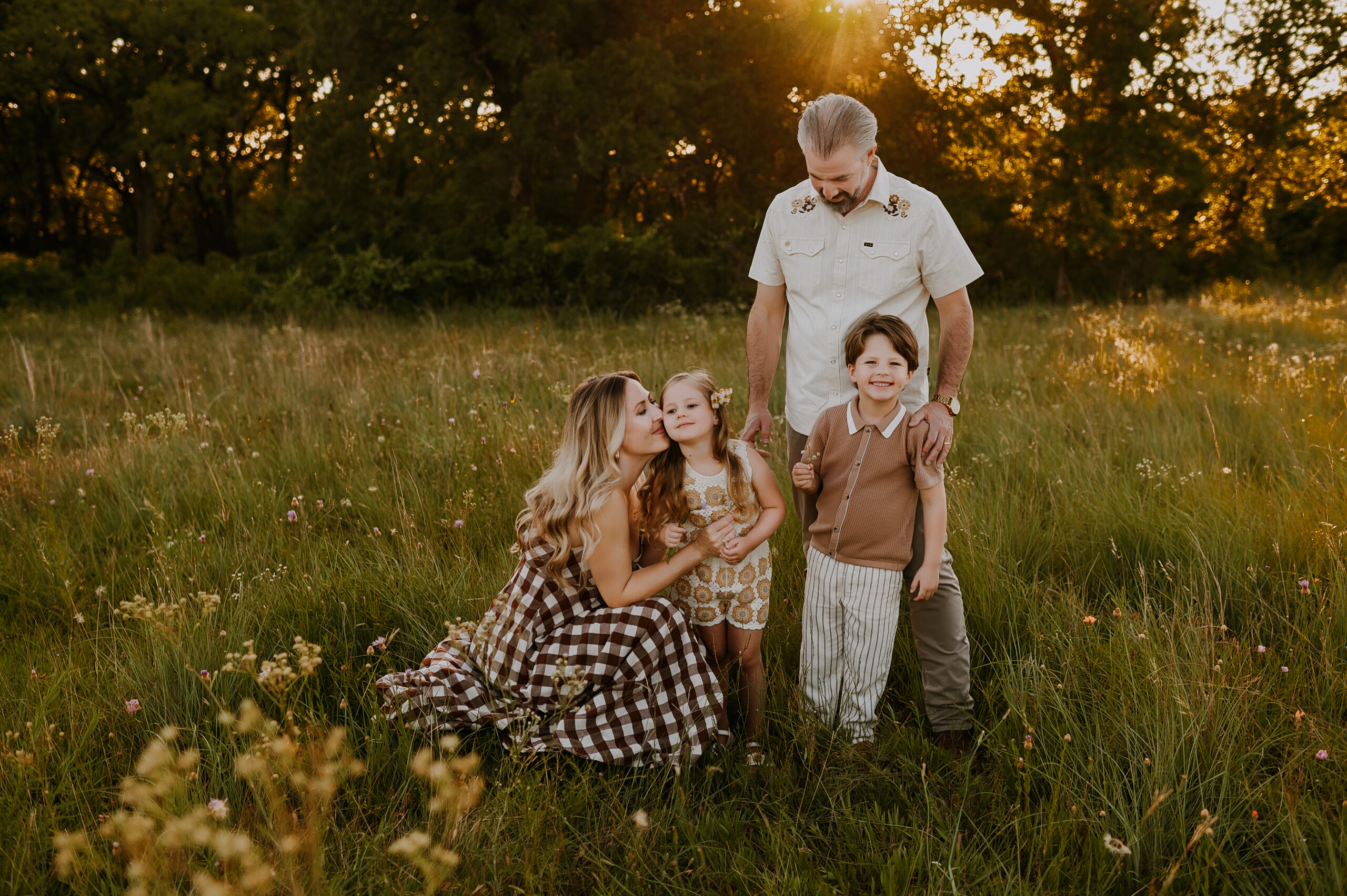 A family of four stand in a golden green and yellow field in fort worth texas in boho clothing and smile with one another
