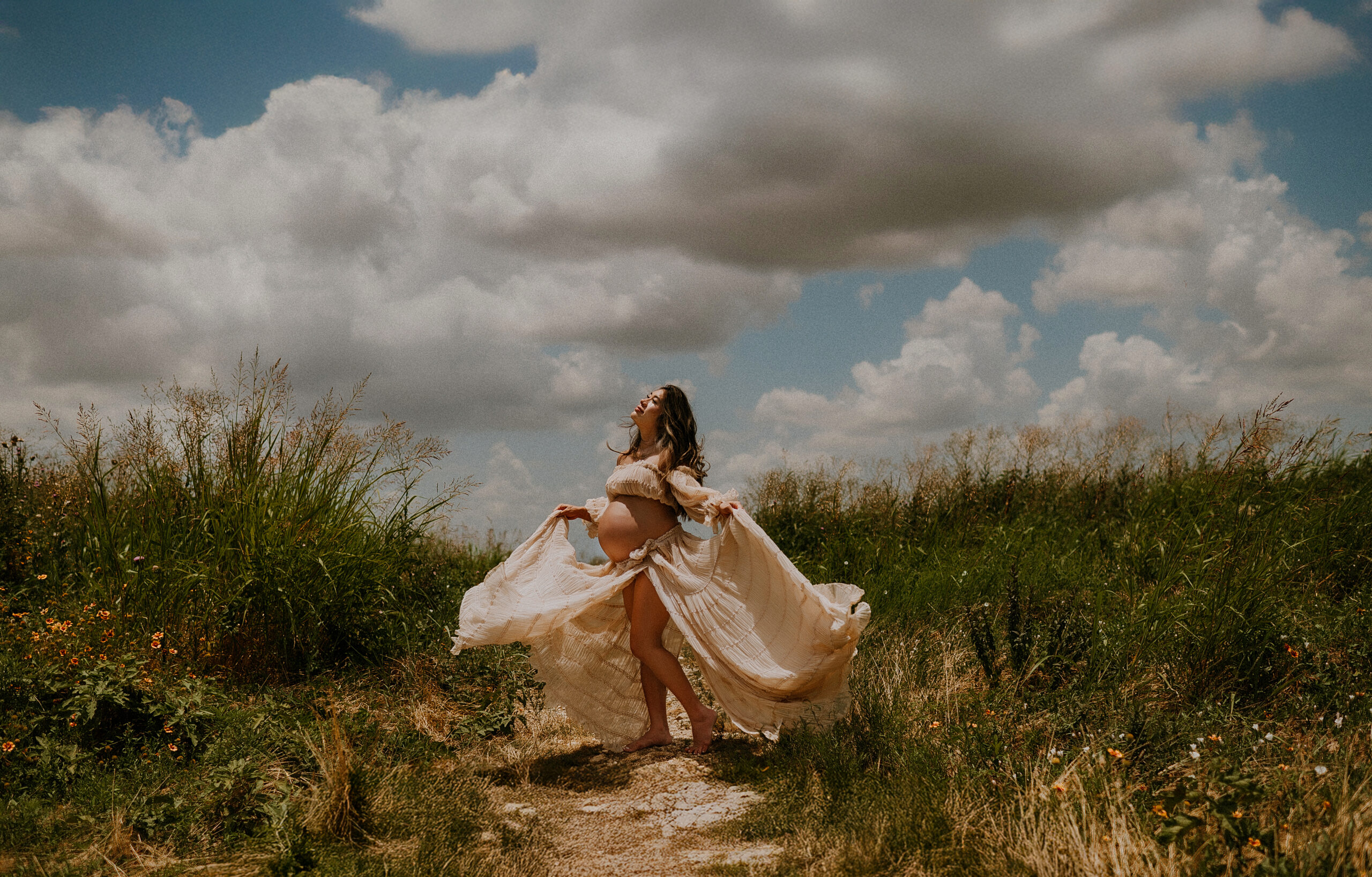 Pregnant woman in flowing cream gown holding her belly under dramatic clouds, photographed by DFW maternity photographer in a wildflower field near Dallas.