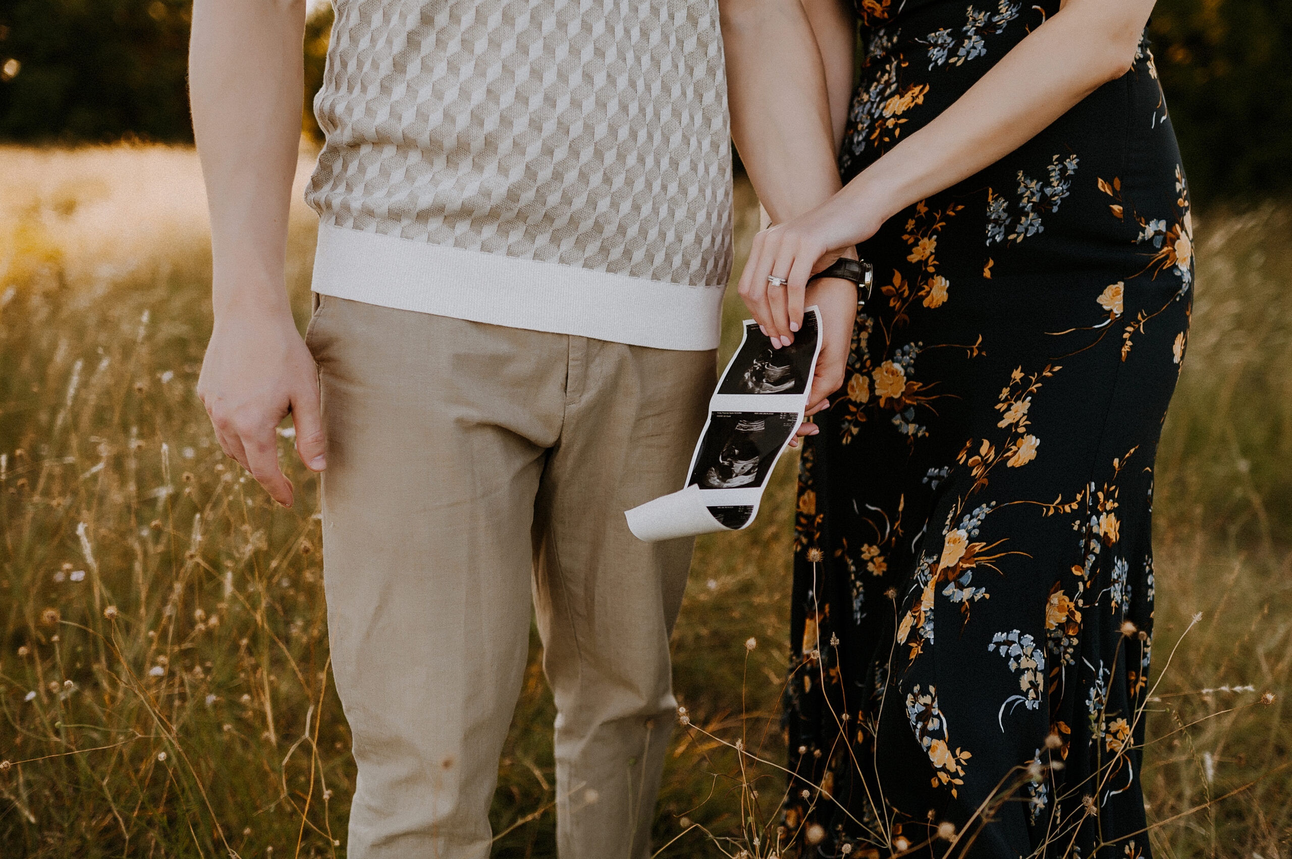 Plano maternity photographer captures pregnancy announcement with couple holding ultrasound photo in golden field at Arbor Hills.