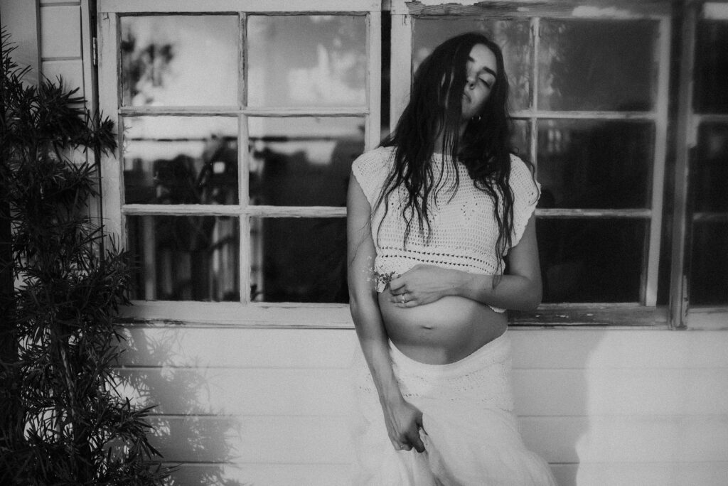 A mom standing against a glass green house and holding flowers to her belly in dallas, texas