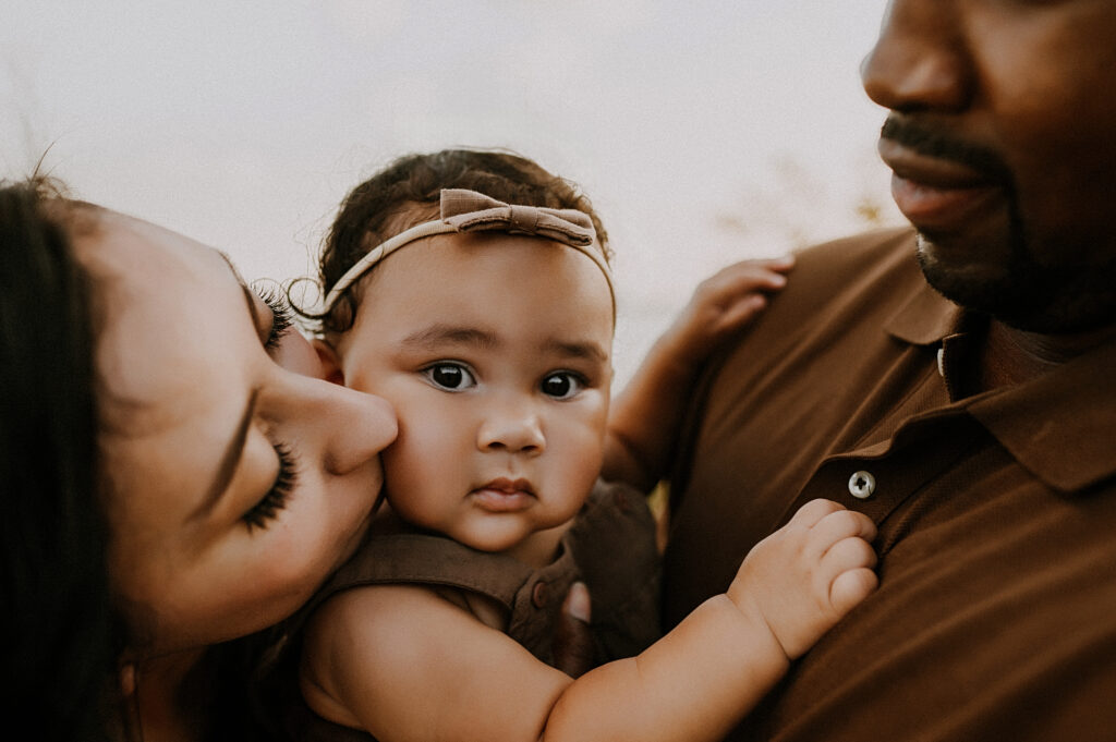 mom is leaning over to kiss her chubby cheeked baby on the cheek while dad holds her in flower mound, texas