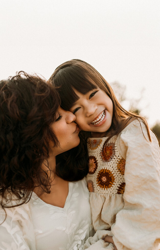 A mom is holding her daughter and kissing her on the cheek as the girl smiles widely at Murrell Park in Flower Mound, TX